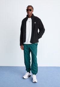 Zwarte fleece jack met hoge kraag, wit T-shirt eronder, groene trainingsbroek en witte sneakers. Accessoires zijn onder andere een paar bruine zonnebrillen.