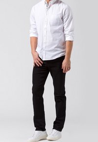 Camisa blanca de manga larga con botones y bolsillo en el pecho, combinada con pantalones slim-fit negros y zapatillas blancas. Diseño simple y limpio.