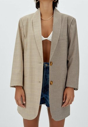 Vrouw draagt oversized blazer met twee verschillende geruite patronen, witte bikini top en denim shorts tegen een effen achtergrond.
