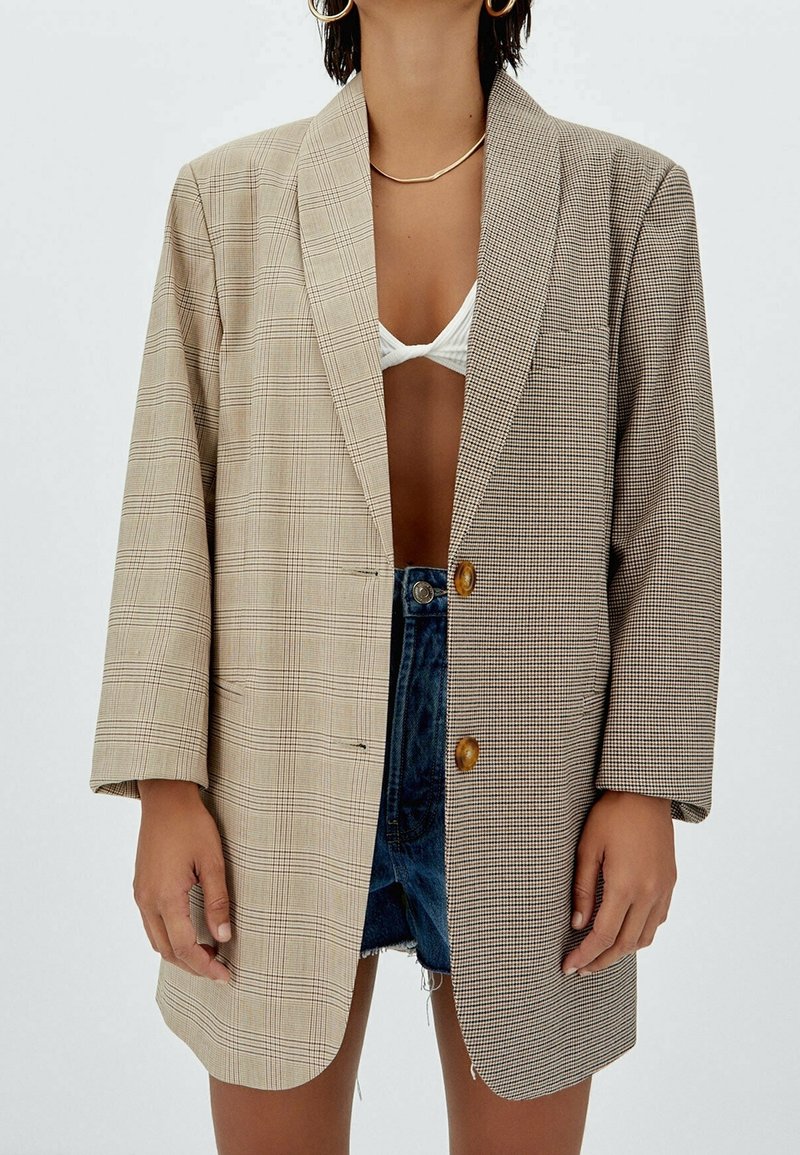 Femme portant un blazer oversize avec deux motifs à carreaux différents, un haut de bikini blanc et un short en jean sur fond uni.