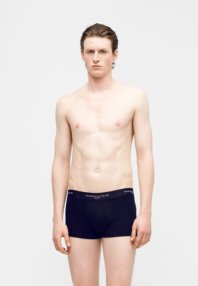 Νέος άνδρας με ανοιχτόχρωμο δέρμα και καστανά μαλλιά που φοράει μαύρα εσώρουχα Maison Kitsuné, στέκεται μπροστά σε απλό λευκό φόντο.