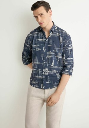 Mężczyzna ubrany w granatową koszulę zapinaną na guziki z białymi wzorami żaglówek oraz jasnobeżowe spodnie, stojący na białym tle.