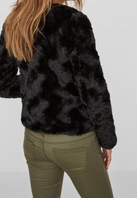 Zwarte nepbontjas met een gestructureerd, golvend oppervlak en een ronde kraag, gecombineerd met olijfgroene aansluitende broek, gezien van de achterkant.