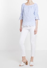 Light blue striped blouse with three-quarter puff sleeves, embroidered scalloped hem, paired with white fitted pants and heeled mules.