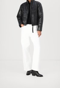 Czarna pikowana kurtka z wysokim kołnierzem, zestawiona z luźnymi białymi spodniami i czarnymi botkami na obcasie, na jednolitym jasnym tle.