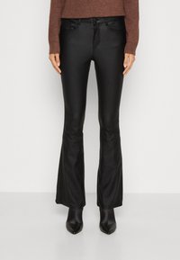 Pantalon noir évasé en matériau élégant et brillant avec une taille ajustée. Il comprend deux poches avant et une fermeture éclair, associé à des talons pointus noirs.