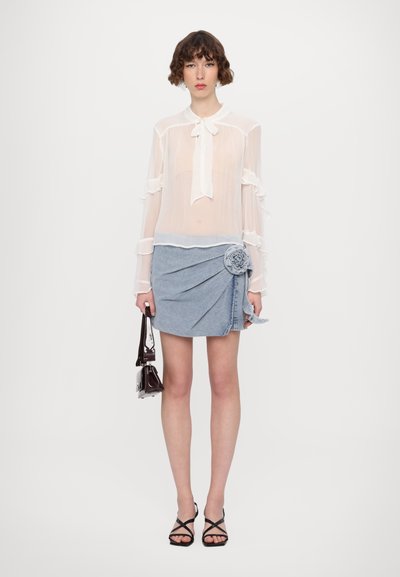 Gennemsigtig hvid bluse med flæser på ærmerne og bindebånd; denim miniskørt med blomstertilføjelse; model der holder en lille mørk håndtaske.