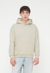 Φωτεινή μπεζ φούτερ με κουκούλα και τσέπη καγκουρό, κορδόνι στη κουκούλα και ριμπ μανσέτες. Το ύφασμα φαίνεται απαλό και ελαφρώς υφασμένο. Φορεμένο με ανοιχτόχρωμο μπλε τζιν.