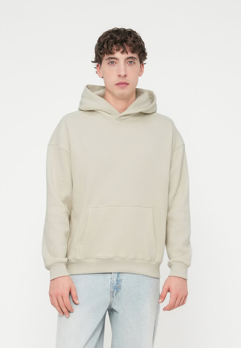 Φωτεινή μπεζ φούτερ με κουκούλα και τσέπη καγκουρό, κορδόνι στη κουκούλα και ριμπ μανσέτες. Το ύφασμα φαίνεται απαλό και ελαφρώς υφασμένο. Φορεμένο με ανοιχτόχρωμο μπλε τζιν.