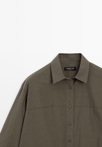 Olive green shirt with a collared neckline, front buttons, and a smooth, textured finish. Features a horizontal seam across the chest.