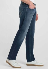 Jeans en denim bleu foncé avec une coupe droite, dotés d'un style classique à cinq poches et d'une légère décoloration. Associés à des baskets blanches.