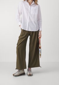 Persona che indossa una camicia bianca a maniche lunghe, pantaloni larghi olive, scarpe beige, mentre tiene una piccola borsa a righe multicolori al lato.