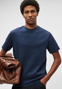 Tumši zila īsām piedurknēm T-krekls no mīksta auduma ar ribotu apaļu kakla izgriezumu un nelielu izšūtu logotipu. Modelis tur brūnu ādas somu.