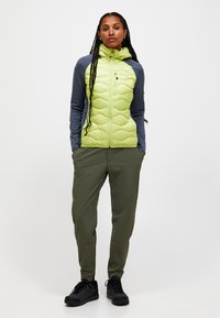Lys grøn puffjakke med quiltet mønster, grå ærmer og fuld lynlås. Olivengrønne bukser og sorte sko fuldender outfittet.