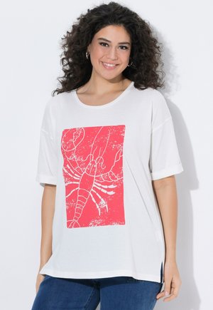 Lächelnde Frau mit lockigem Haar, die ein weißes T-Shirt mit großem rotem Hummeraufdruck trägt, kombiniert mit blauen Jeans.