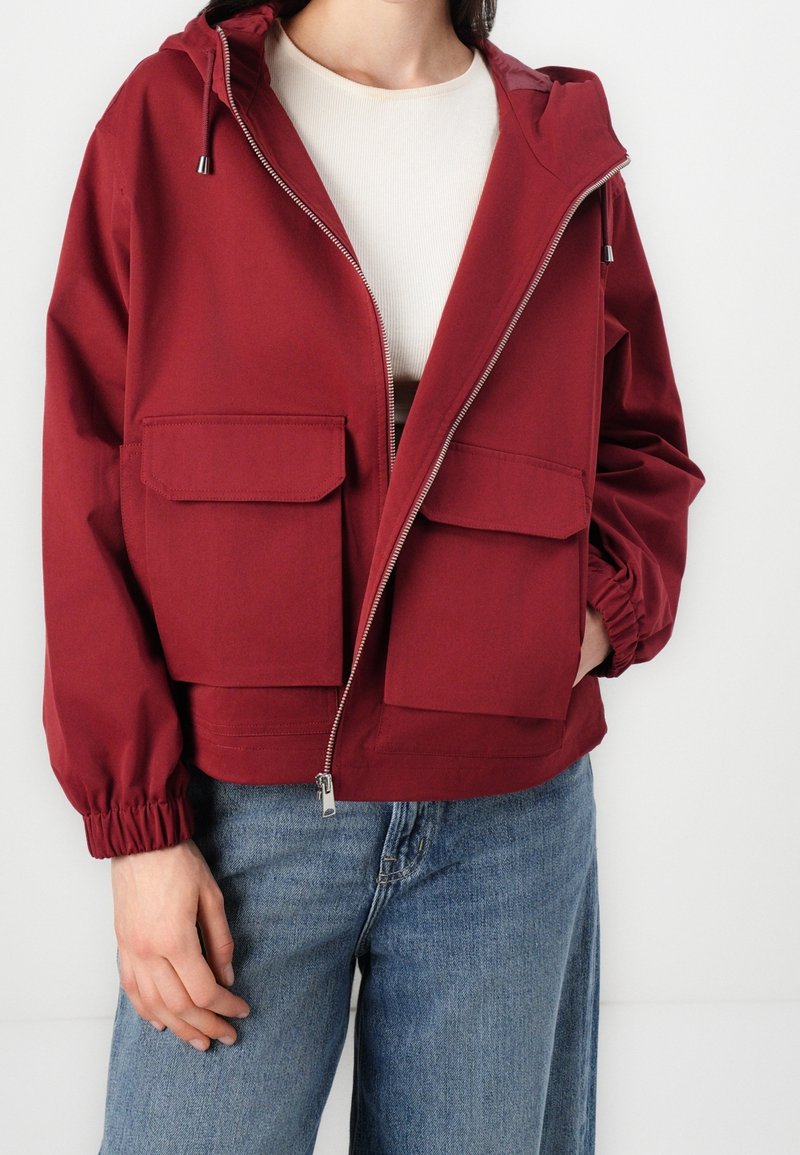 Personne portant une veste zippée bordeaux avec de grandes poches avant et des poignets élastiques, associée à un jean bleu et un haut blanc.