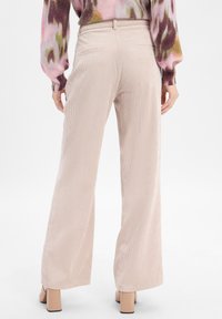 Persoon in beige wijde corduroy broek, langharig shirt met een multikleurig patroon, en beige blokhakken, staande met de rug naar de camera.