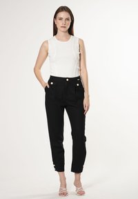 Top blanco sin mangas con líneas verticales texturizadas. Pantalones negros de alta cintura con dos bolsillos frontales y acentos de botones dorados. Fondo claro.