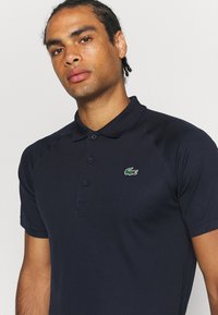 Polo de color azul marino con un cuello clásico, tres botones y el logo de cocodrilo verde de Lacoste en el lado izquierdo del pecho. Fabricado con un tejido liso y texturizado.
