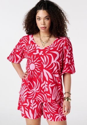 Femme aux cheveux bouclés porte un haut à manches courtes imprimé tropical rouge et rose avec un short assorti, accessoirisée de bijoux en or, posant avec assurance.