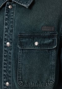 Tummansininen farkkutakki, jossa on tekstuuripinta, etutasku, hopeiset yksityiskohdat ja musta brändilabel vasemmassa rinnassa.