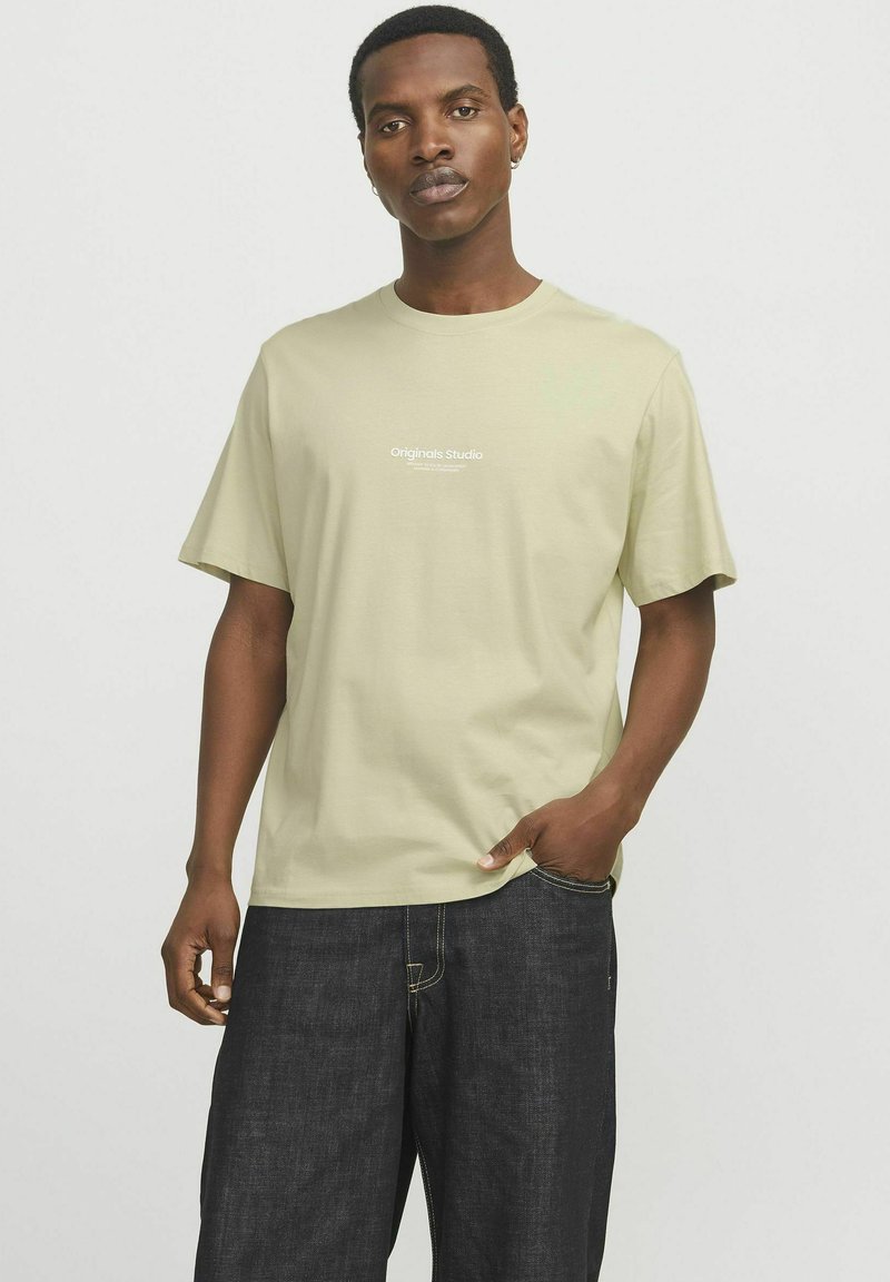 Hellgrünes Baumwoll-T-Shirt mit kurzen Ärmeln, rundem Halsausschnitt und weißem "Originals Studio"-Schriftzug auf der Brust. Getragen mit dunklen Jeans.