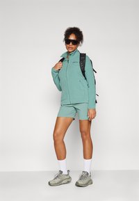 Veste en polaire teal clair avec un zip à l'avant, associée à un short assorti. Sac à dos noir, chaussettes blanches et chaussures de sport grises.