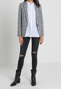 Šedý kostičkovaný blazer se strukturovaným střihem, nošený přes bílou košili s knoflíky. Doplněno černými potrhanými úzkými jeany a černými kotníkovými botami.