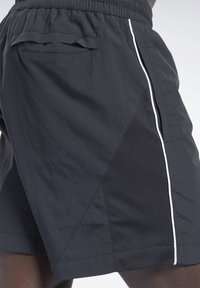 Shorts de sport noirs avec une ceinture élastique, une poche arrière à rabat, un panneau en mesh latéral et un détail de rayure verticale blanche.