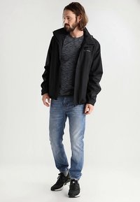 Mann med skulderlangt hår og skjegg som har på seg en svart jakke, mørkegrå skjorte, blå jeans og svarte joggesko, stående mot en hvit bakgrunn.