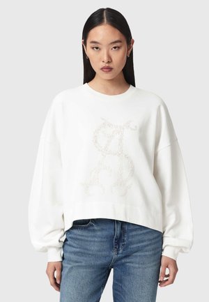 Frau mit langen, glatten Haaren, die einen lockeren weißen Pullover mit einem strukturierten Monogramm und blaue Jeans trägt, steht vor einem einfarbigen Hintergrund.
