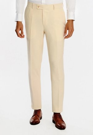 Mężczyzna w beżowych, dopasowanych spodniach i brązowych, skórzanych eleganckich butach, stoi z rękami swobodnie opuszczonymi wzdłuż ciała.