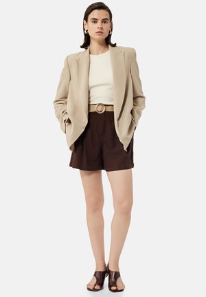 Kvinde iført beige blazer, hvid top, brune bælte-shorts og brune åbne hæle, står med hænderne i lommerne mod en ensfarvet baggrund.