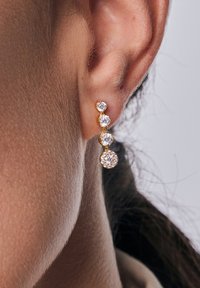 Złote kolczyki zdobią cztery stopniowo rosnące, przezroczyste kryształowe kamienie, z największym kamieniem na dole, tworząc efekt kaskady.