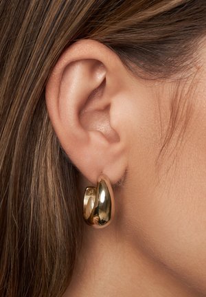 Boucle d'oreille en or présentée sur un lobe, avec une surface lisse et réfléchissante et une forme arrondie avec une légère ouverture.