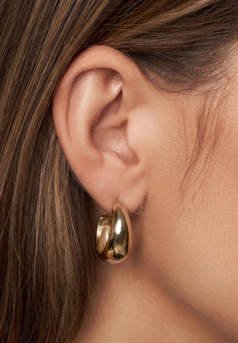 Boucle d'oreille en or présentée sur un lobe, avec une surface lisse et réfléchissante et une forme arrondie avec une légère ouverture.