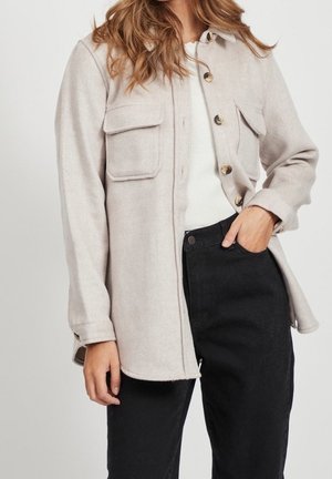 Mujer con chaqueta tipo camisa color beige claro con botones y bolsillos con solapa sobre una camiseta blanca y pantalones negros, con la mano en el bolsillo, de pie contra un fondo blanco.