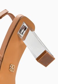 Sandalia de tacón alto en color beige con un tacón bloque transparente. Cuenta con una parte superior suave y mate, correa ajustable y un detalle dorado en la suela.