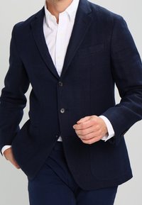 Man som har på sig en marinblå kavaj över en vit skjorta med knappar, med matchande marinblå byxor, och justerar sin jacka.