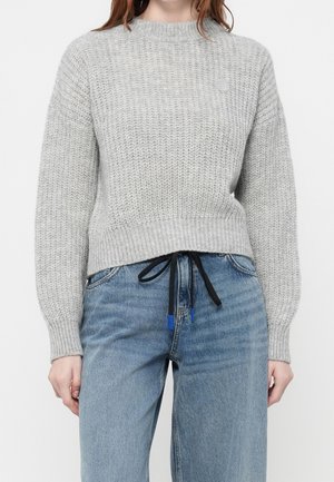 Gros plan d’une personne portant un pull en maille gris clair à manches longues et un jean bleu avec un cordon noir noué à la taille.
