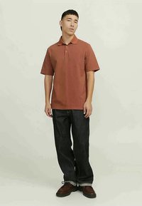 Πολό φανέλα σε χαλκό χρώμα με γιακά, κοντά μανίκια και άνετη εφαρμογή, συνδυασμένη με σκούρα μπλε, φαρδιά τζην και καφέ παπούτσια.