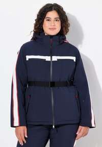 Námořnická modrá zateplená bunda s bílými a červenými pruhy, má zipový přední díl, dvě náprsní kapsy, nastavovací kapuci a opasek v pase.