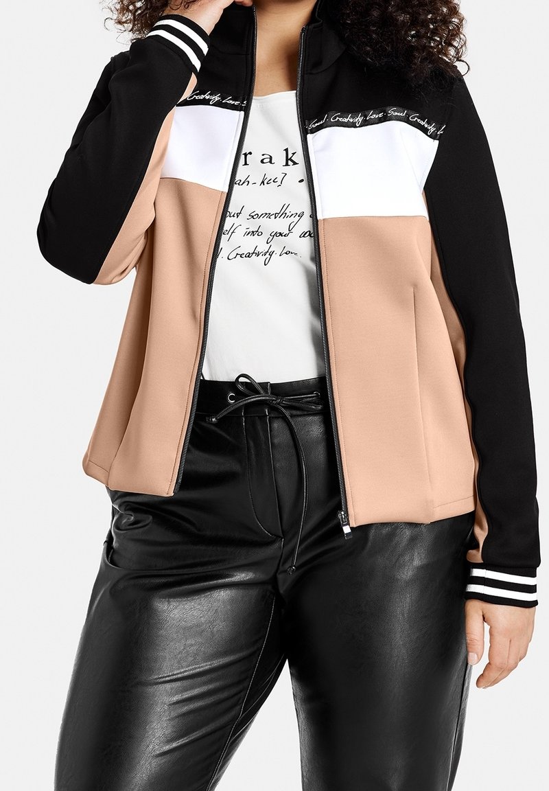 Femme portant une veste zippée aux panneaux noirs, blancs et beiges, un pantalon en cuir noir et un t-shirt blanc imprimé en dessous.