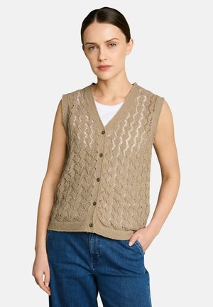 Woman wearing a sleeveless beige knit buttoned vest over a white top with blue jeans, standing with one hand in pocket against white background.