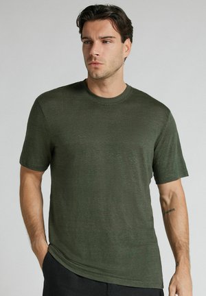 Hombre con cabello oscuro que lleva una camiseta simple de manga corta y cuello redondo de color verde oliva, de pie con una mano en el bolsillo, mirando hacia un lado.