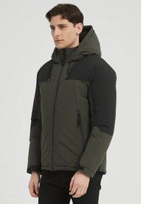 Veste isolante verte et noire avec capuche, dotée d'une fermeture éclair, de cordons de serrage réglables et de poches latérales zippées.