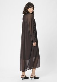 Žena v dlhých priesvitných hnedých šatách s volánovými rukávmi, s čiernymi slingback kotníkovými podpätkami, stojí a pozerá cez svoje rameno.