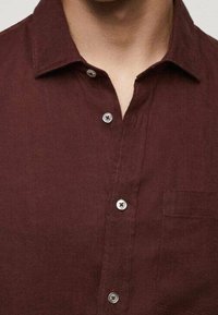 Nahaufnahme einer Person in einem dunkelroten Hemd mit Knöpfen und einem Kragen, das die sichtbaren Knöpfe an der Vorderseite zeigt.