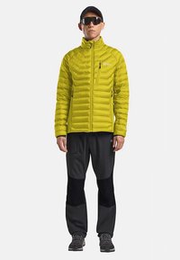 Gelbe isolierte Jacke mit gesteppter Optik und Reißverschlusstaschen, kombiniert mit schwarzen Hosen mit strukturierten Einsätzen und seitlichem Reißverschluss. Sonnenbrille und Mütze werden getragen.