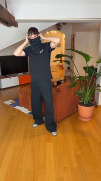 Melns kapuces apģērbs, kas turēts augšup, savienots ar melnu T-kreklu un platām melnām biksēm. Telpā redzama koka grīda un tuvumā esošs podos augs.